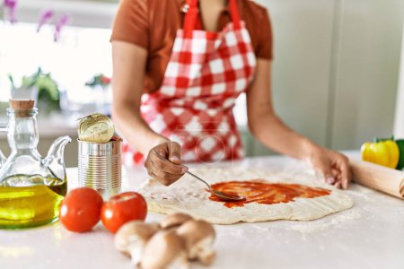 Téléchargez les photos : Young beautiful hispanic woman putting tomato sauce on dough pizza at the kitchen - en image libre de droit