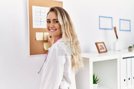Photo for Young woman wearing doctor uniform writing on corkboard at clinic - Royalty Free Image