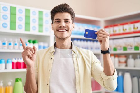 Téléchargez les photos : Jeune homme hispanique à la pharmacie pharmacie tenant carte de crédit souriant heureux pointant avec la main et le doigt sur le côté - en image libre de droit