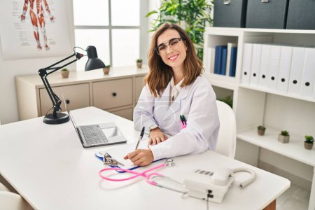 Photo for Young woman wearing doctor uniform writing medical report at clinic - Royalty Free Image