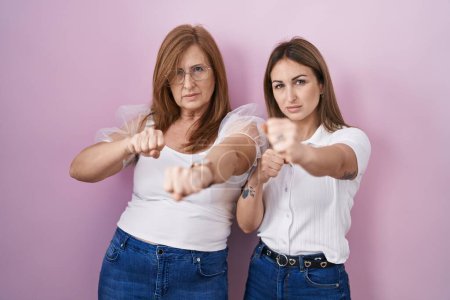 Foto de Madre e hija hispanas con camiseta blanca casual sobre fondo rosa golpeando puño para luchar, ataque agresivo y enojado, amenaza y violencia - Imagen libre de derechos