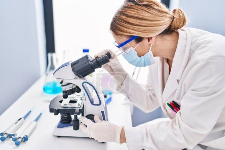 Téléchargez les photos : Young blonde woman scientist wearing medical mask using microscope at laboratory - en image libre de droit