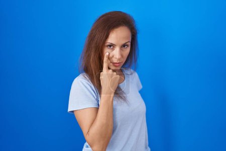 Téléchargez les photos : Brunette femme debout sur fond bleu pointant vers l'oeil vous regarder geste, expression suspecte - en image libre de droit