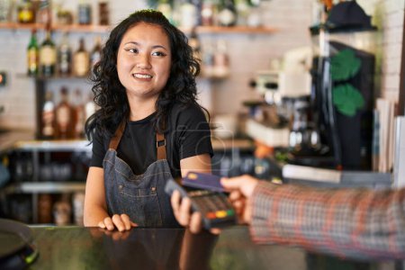 Foto de Joven camarera china sonriendo confiada usando tarjeta de crédito y teléfono de datos en el restaurante - Imagen libre de derechos