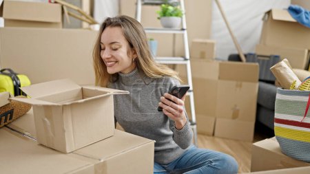 Photo for Young caucasian woman unpacking cardboard box using smartphone at new home - Royalty Free Image