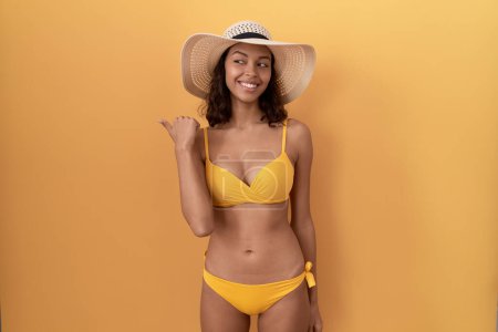 Téléchargez les photos : Jeune femme hispanique portant un bikini et un chapeau d'été souriant avec un visage heureux regardant et pointant vers le côté avec le pouce vers le haut. - en image libre de droit