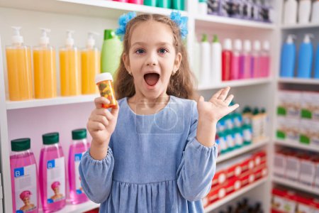Téléchargez les photos : Jeune fille tenant des pilules à la pharmacie célébrant la victoire avec un sourire heureux et l'expression gagnante avec les mains levées - en image libre de droit