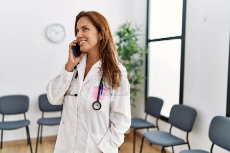 Photo for Young latin woman wearing doctor uniform talking on the smartphone at clinic waiting room - Royalty Free Image