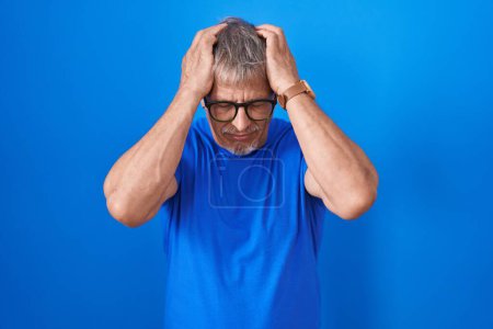 Téléchargez les photos : Homme hispanique avec les cheveux gris debout sur fond bleu souffrant de maux de tête désespérés et stressés parce que la douleur et la migraine. mains sur la tête. - en image libre de droit