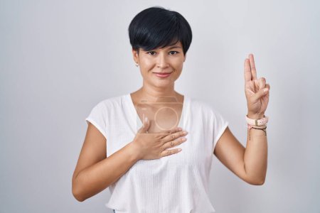 Téléchargez les photos : Jeune femme asiatique avec les cheveux courts debout sur fond isolé souriant jurer avec la main sur la poitrine et les doigts vers le haut, ce qui rend un serment de fidélité promesse - en image libre de droit