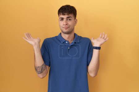 Photo for Young hispanic man standing over yellow background clueless and confused expression with arms and hands raised. doubt concept. - Royalty Free Image
