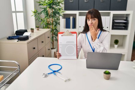 Photo for Young brunette doctor woman holding clipboard covering mouth with hand, shocked and afraid for mistake. surprised expression - Royalty Free Image