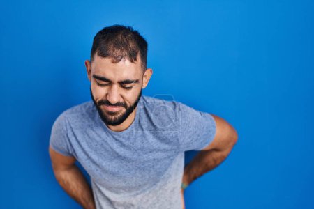 Téléchargez les photos : Moyen-Orient homme avec barbe debout sur fond bleu souffrant de maux de dos, toucher le dos avec la main, douleurs musculaires - en image libre de droit
