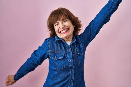 Téléchargez les photos : Femme d'âge moyen debout sur fond rose dansant heureux et joyeux, souriant décontracté et confiant écoutant de la musique - en image libre de droit