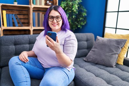 Téléchargez les photos : Jeune belle femme plus la taille en utilisant smartphone assis sur le canapé à la maison - en image libre de droit