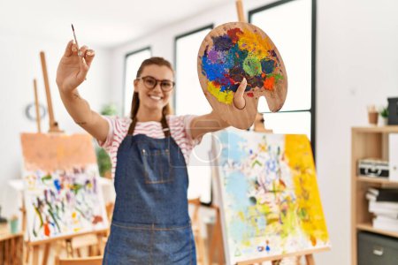 Photo for Young woman smiling confident holding paintbrush and palette at art studio - Royalty Free Image