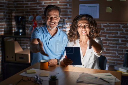 Photo for Middle age hispanic couple using touchpad sitting on the table at night smiling friendly offering handshake as greeting and welcoming. successful business. - Royalty Free Image