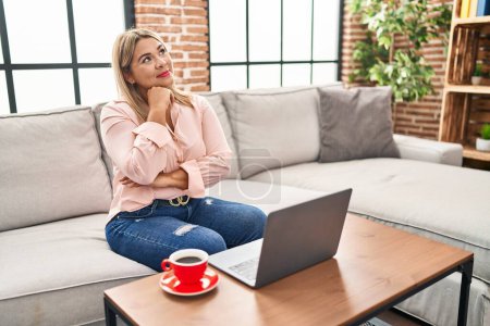 Photo for Young hispanic woman using laptop sitting on the sofa at home with hand on chin thinking about question, pensive expression. smiling with thoughtful face. doubt concept. - Royalty Free Image