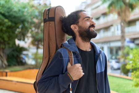 Foto de Young hispanic man musician smiling confident holding guitar case at park - Imagen libre de derechos