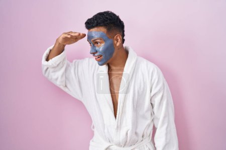 Photo for Young hispanic man wearing beauty face mask and bath robe very happy and smiling looking far away with hand over head. searching concept. - Royalty Free Image