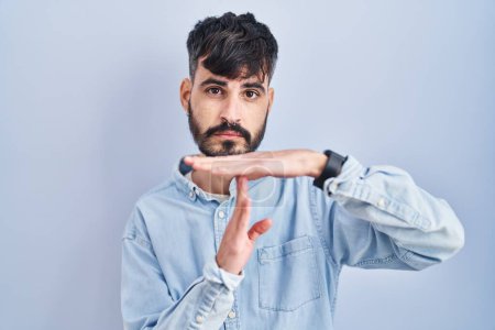 Foto de Joven hombre hispano con barba de pie sobre fondo azul haciendo un gesto de tiempo fuera con las manos, rostro frustrado y serio - Imagen libre de derechos