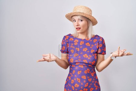 Téléchargez les photos : Jeune femme caucasienne portant une robe de fleurs et un chapeau d'été désemparé et confus à bras ouverts, aucun concept d'idée. - en image libre de droit