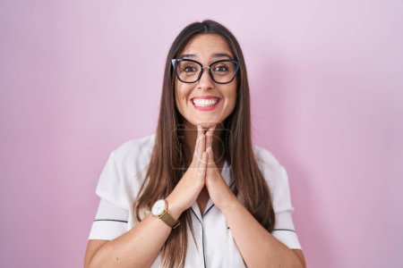 Photo for Young brunette woman wearing glasses standing over pink background praying with hands together asking for forgiveness smiling confident. - Royalty Free Image