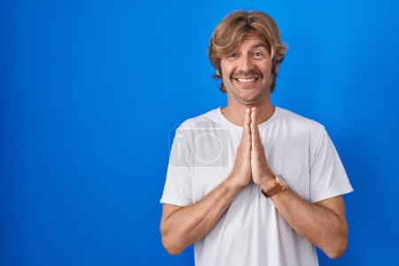 Photo for Middle age man standing over blue background praying with hands together asking for forgiveness smiling confident. - Royalty Free Image
