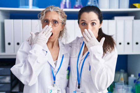 Foto de Madre e hija trabajando en el laboratorio científico cubriendo la boca con la mano, conmocionadas y temerosas de equivocarse. expresión sorprendida - Imagen libre de derechos