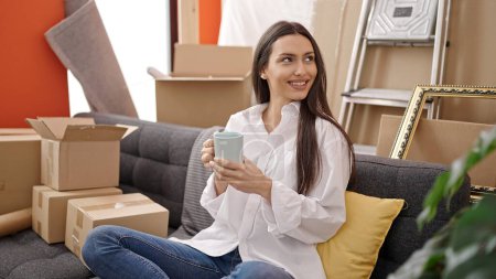 Photo for Young beautiful hispanic woman drinking coffee sitting on sofa at new home - Royalty Free Image