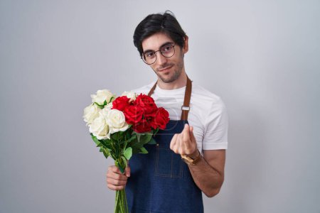 Photo for Young hispanic man holding bouquet of white and red roses doing money gesture with hands, asking for salary payment, millionaire business - Royalty Free Image
