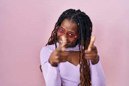 Foto de Mujer africana con el pelo trenzado de pie sobre el fondo rosa señalando los dedos a la cámara con la cara feliz y divertida. buena energía y vibraciones. - Imagen libre de derechos