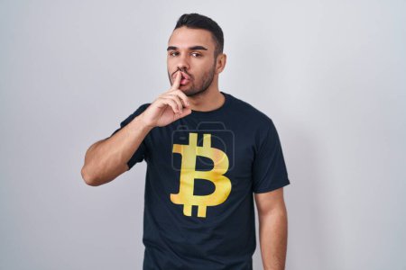Téléchargez les photos : Jeune homme hispanique portant Bitcoin t-shirt demandant à être calme avec le doigt sur les lèvres. silence et concept secret. - en image libre de droit