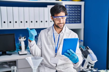 Foto de Hombre latino guapo trabajando en el laboratorio científico usando la tableta apuntando el pulgar hacia el lado sonriendo feliz con la boca abierta - Imagen libre de derechos