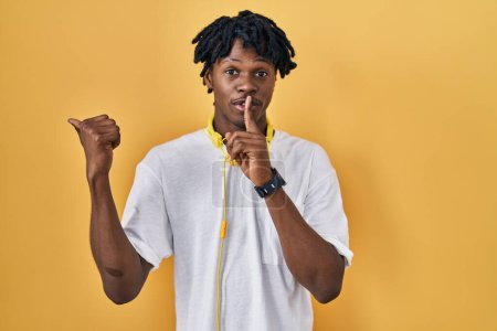 Téléchargez les photos : Jeune homme africain avec dreadlocks debout sur fond jaune demandant à être calme avec le doigt sur les lèvres pointant avec la main sur le côté. silence et concept secret. - en image libre de droit