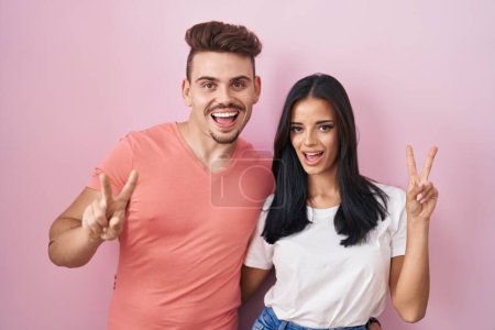 Téléchargez les photos : Jeune couple hispanique debout sur fond rose souriant avec un sourire heureux clin d'oeil à la caméra faisant signe de victoire avec les doigts. numéro deux. - en image libre de droit