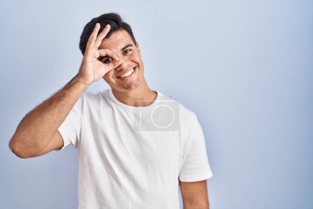 Téléchargez les photos : Homme hispanique debout sur fond bleu faisant un geste correct avec la main souriante, les yeux regardant à travers les doigts avec un visage heureux. - en image libre de droit