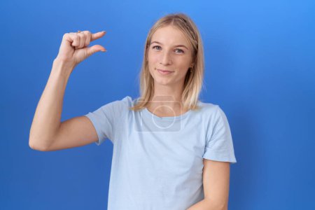 Téléchargez les photos : Jeune femme caucasienne portant casual t-shirt bleu geste souriant et confiant avec la main faisant signe de petite taille avec les doigts à la recherche et la caméra. concept de mesure. - en image libre de droit
