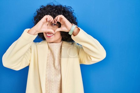 Photo for Young brunette woman with curly hair standing over blue background doing heart shape with hand and fingers smiling looking through sign - Royalty Free Image