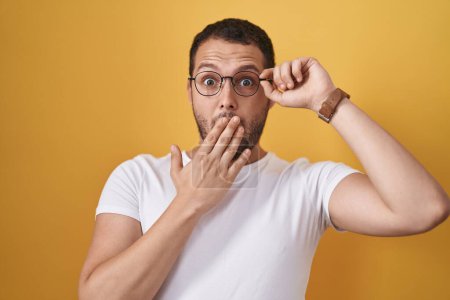 Foto de Hombre hispano sosteniendo gafas cubriendo la boca con la mano, sorprendido y temeroso de equivocarse. expresión sorprendida - Imagen libre de derechos