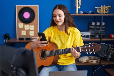 Photo for Young woman musician playing classical guitar using smartphone at music studio - Royalty Free Image