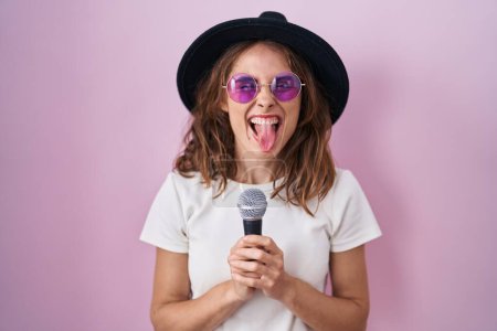 Téléchargez les photos : Belle femme brune chantant chanson à l'aide d'un microphone collant la langue heureux avec une expression drôle. - en image libre de droit