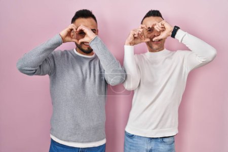 Photo for Homosexual couple standing over pink background doing heart shape with hand and fingers smiling looking through sign - Royalty Free Image