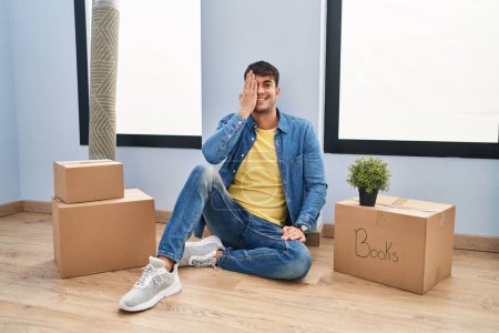 Photo for Young hispanic man sitting on the floor at new home covering one eye with hand, confident smile on face and surprise emotion. - Royalty Free Image