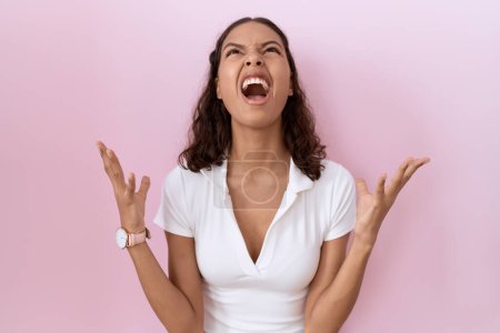 Foto de Mujer hispana joven con camiseta blanca casual loca y loca gritando y gritando con expresión agresiva y los brazos levantados. concepto de frustración. - Imagen libre de derechos