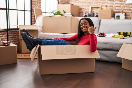 Photo for Young african american with braids moving to a new home inside of a cardboard box pointing thumb up to the side smiling happy with open mouth - Royalty Free Image