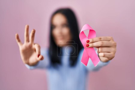 Foto de Mujer hispana sosteniendo cinta de cáncer rosa haciendo signo de ok con los dedos, sonriendo gesto amistoso excelente símbolo - Imagen libre de derechos