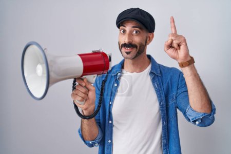 Foto de Hombre hispano con barba gritando a través de megáfono sorprendido con una idea o pregunta apuntando con el dedo con la cara feliz, número uno - Imagen libre de derechos