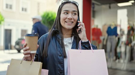 Téléchargez les photos : Jeune belle femme hispanique parlant sur smartphone tenant des sacs à provisions et du café dans la rue - en image libre de droit