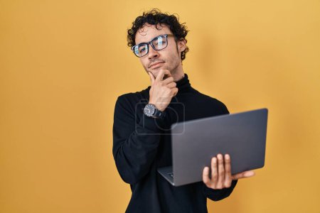 Foto de Hombre hispano trabajando con computadora portátil cara seria pensando en la pregunta con la mano en la barbilla, reflexivo acerca de la idea confusa - Imagen libre de derechos
